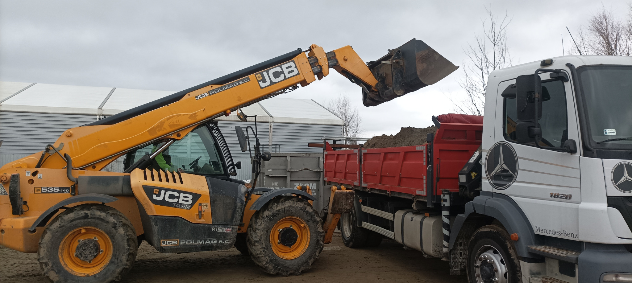Żółta ładowarka teleskopowa JCB ładuje ziemię do czerwonej ciężarówki Mercedes-Benz na placu budowy. Widoczne logo JCB i Mercedes-Benz.