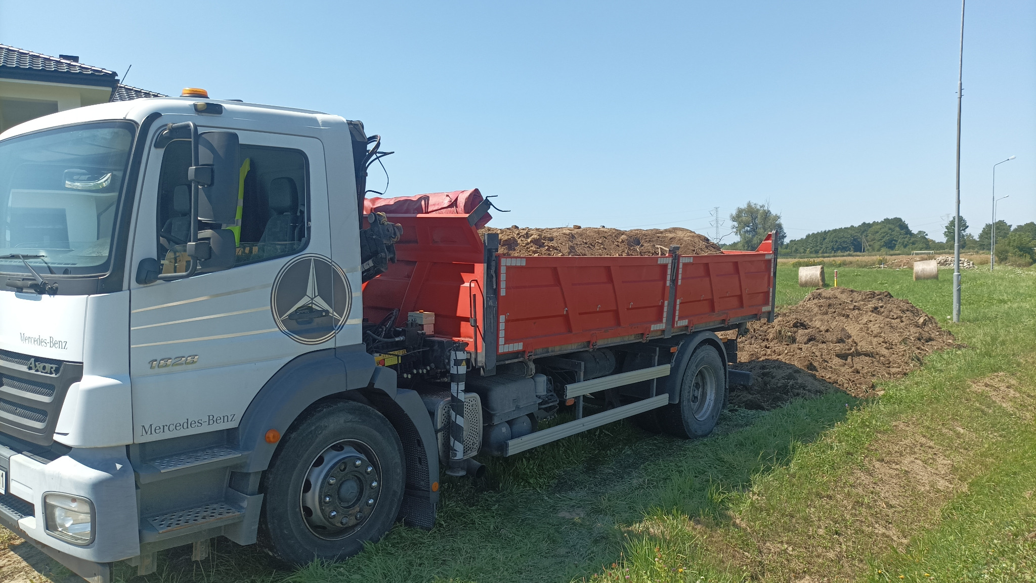 Ciężarówka Mercedes-Benz z czerwoną skrzynią ładunkową wypełnioną ziemią, stojąca na trawiastym terenie w słoneczny dzień. W tle widoczne bele siana i drzewa.