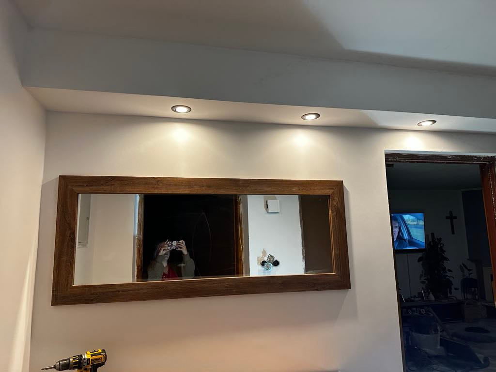 Montaż lustra w drewnianej ramie, widoczne oświetlenie sufitowe i wiertarka. W lustrze odbija się osoba robiąca zdjęcie.