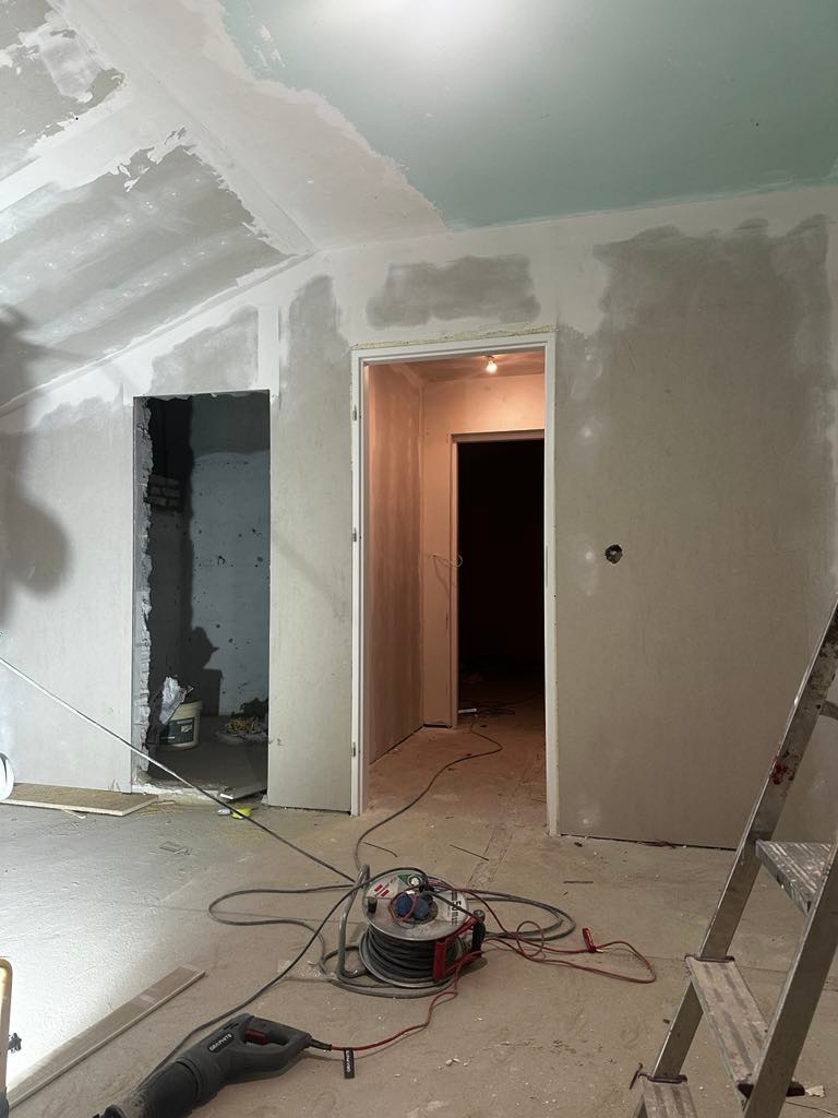 Wnętrze w trakcie prac wykończeniowych: ściany z nałożoną gładzią, widoczne otwory drzwiowe, narzędzia i drabina. Sufit częściowo pomalowany na zielono.