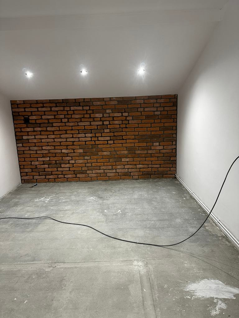 Surowe wnętrze z ceglaną ścianą akcentową i betonową podłogą. Widoczne trzy punktowe źródła światła w suficie. Kabel elektryczny leży na podłodze.