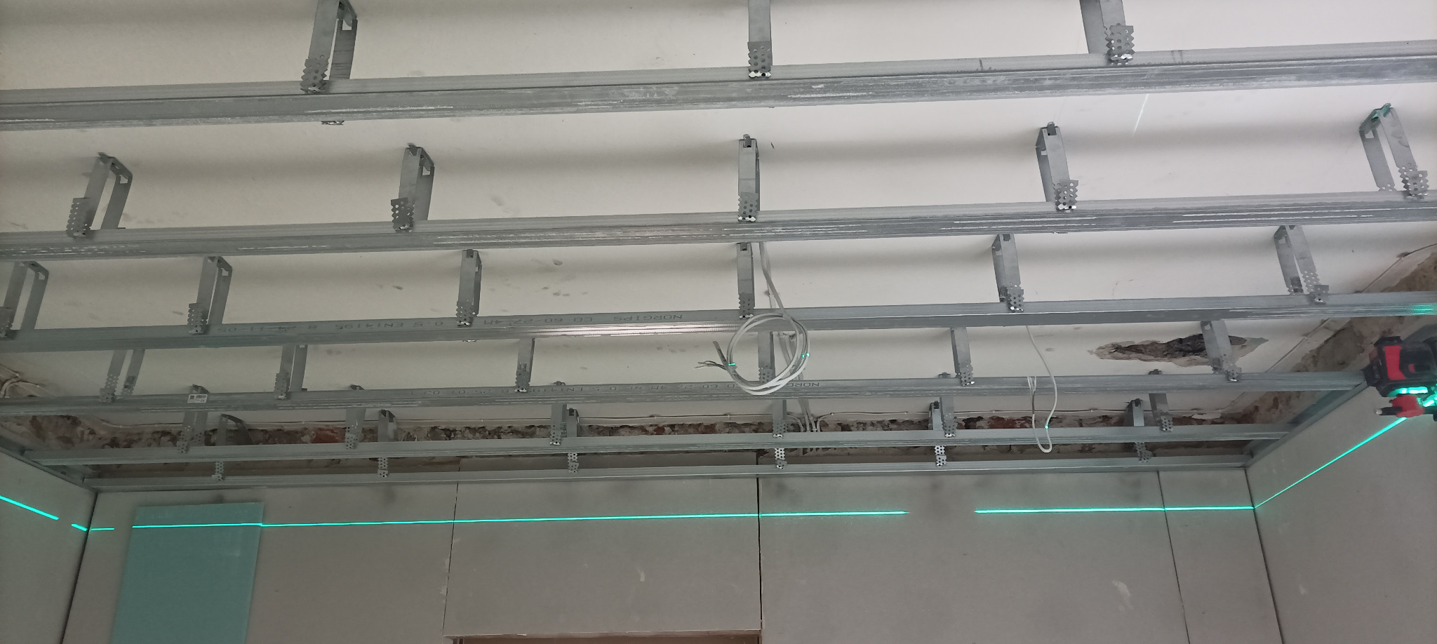 Montaż metalowej konstrukcji sufitu podwieszanego z płyt karton-gips, widoczny laser krzyżowy wyznaczający poziom. Etap prac przed zakryciem instalacji.