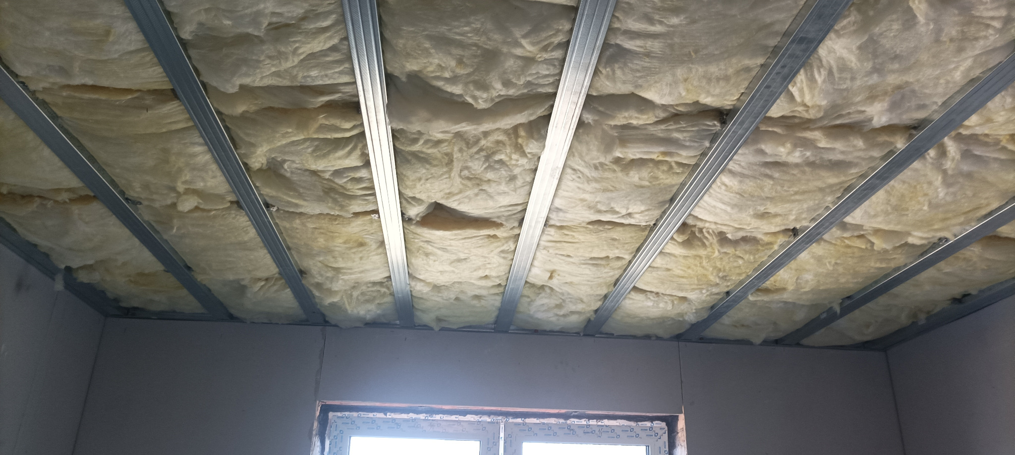 Ocieplenie poddasza wełną mineralną, widoczne metalowe profile konstrukcyjne. Fragment okna w dolnej części kadru. Prace wykończeniowe wewnątrz budynku.