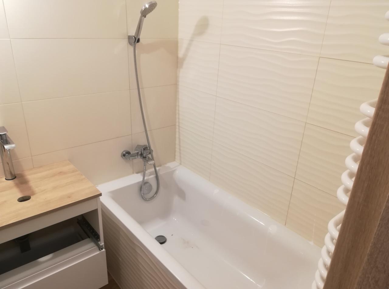 Nowoczesna łazienka z beżowymi płytkami o falistej strukturze, białą wanną, chromowaną baterią i szafką pod umywalkę z blatem imitującym drewno.