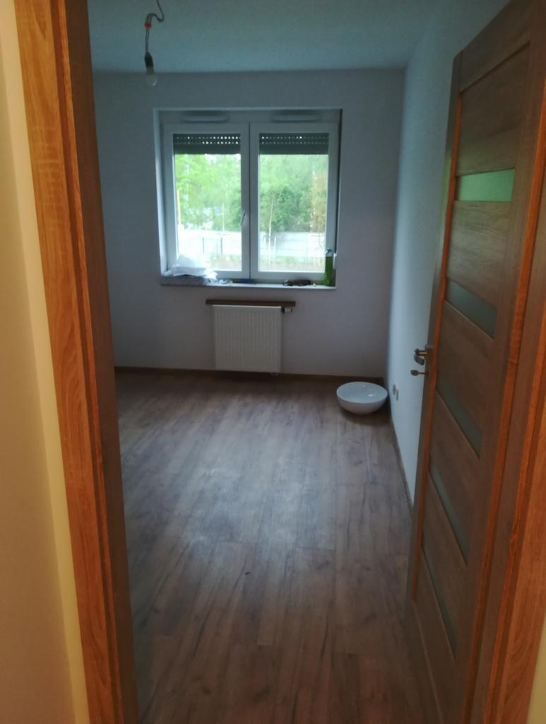 Pokój z drewnopodobną podłogą, białymi ścianami, grzejnikiem pod oknem z roletami i drewnianymi drzwiami z przeszkleniami, widziany z korytarza.