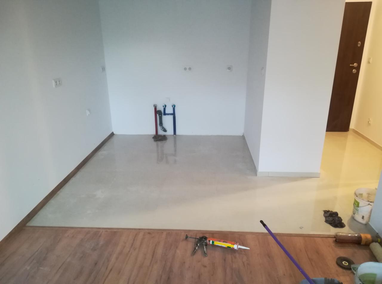 Podłoga w trakcie łączenia dwóch rodzajów powierzchni: drewnopodobnej i jasnych płytek; widoczne narzędzia i materiały montażowe, rury instalacji wodnej wystające ze ściany.