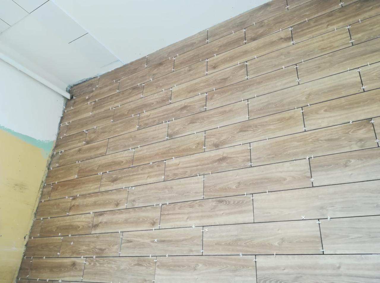 Ściana wyłożona podłużnymi płytkami imitującymi drewno, z białymi krzyżykami dystansowymi w spoinach, widoczny fragment sufitu i fragment niepomalowanej ściany bocznej.