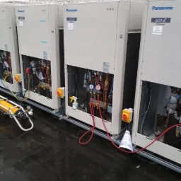 Rząd jednostek zewnętrznych Panasonic ECO i, z otwartymi panelami frontowymi ukazującymi instalacje rurowe, podłączone do manometrów i butli z gazem na wózku, ustawionych na mokrym dachu.