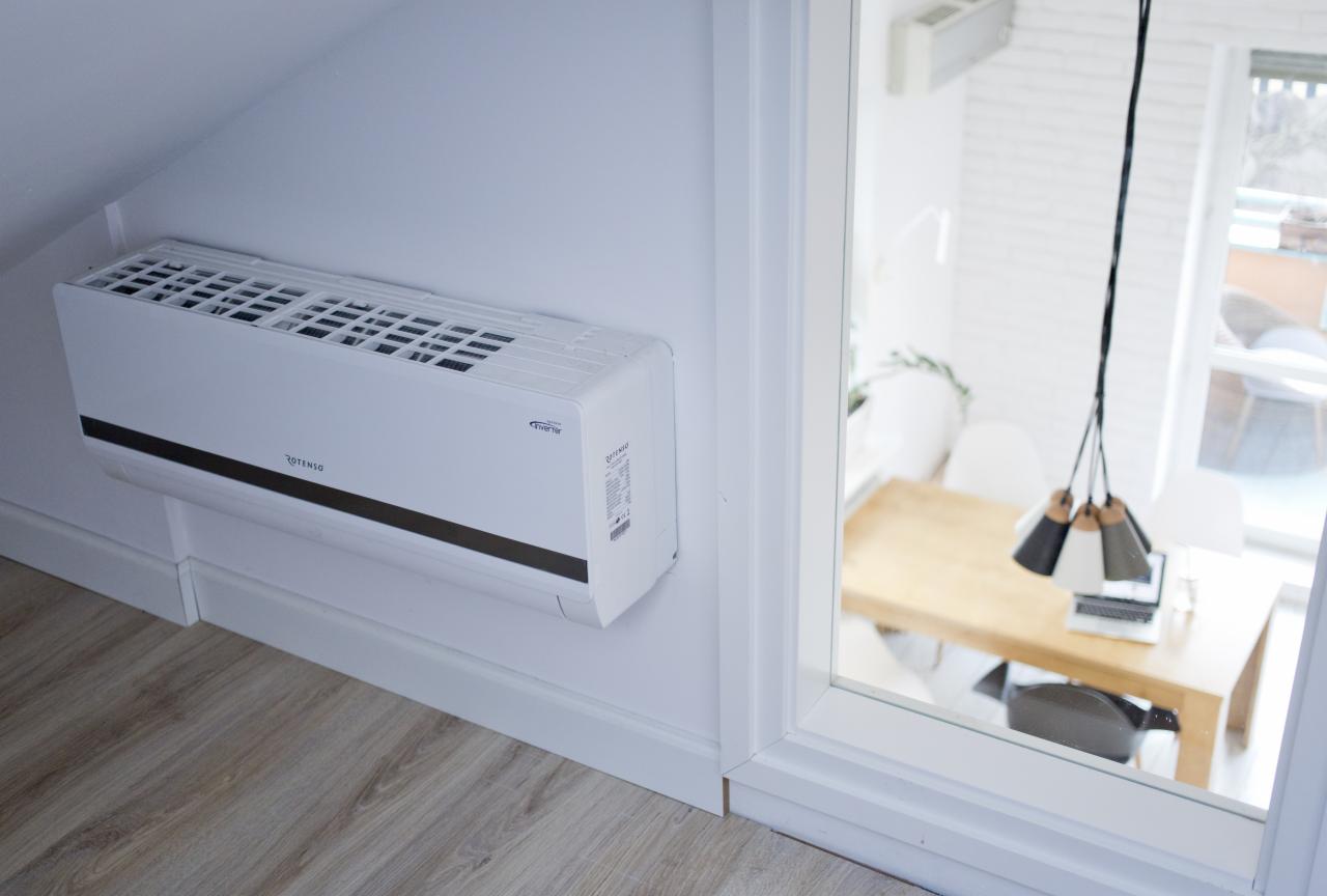 Biała jednostka wewnętrzna klimatyzatora marki Rotenso zamontowana na ścianie w jasnym pomieszczeniu z widokiem na stół i lampę przez okno.