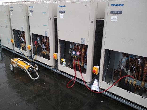 Rząd jednostek zewnętrznych Panasonic ECO i, z otwartymi panelami frontowymi ukazującymi instalacje rurowe, podłączone do manometrów i butli z gazem na wózku, ustawionych na mokrym dachu.