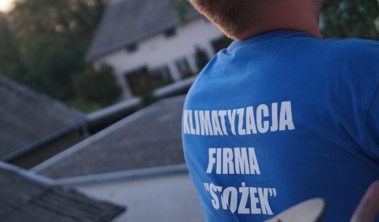 Mężczyzna w niebieskiej koszulce z białym napisem 'KLIMATYZACJA FIRMA 'SÓŻEK'', widok od tyłu, na tle dachów budynków.