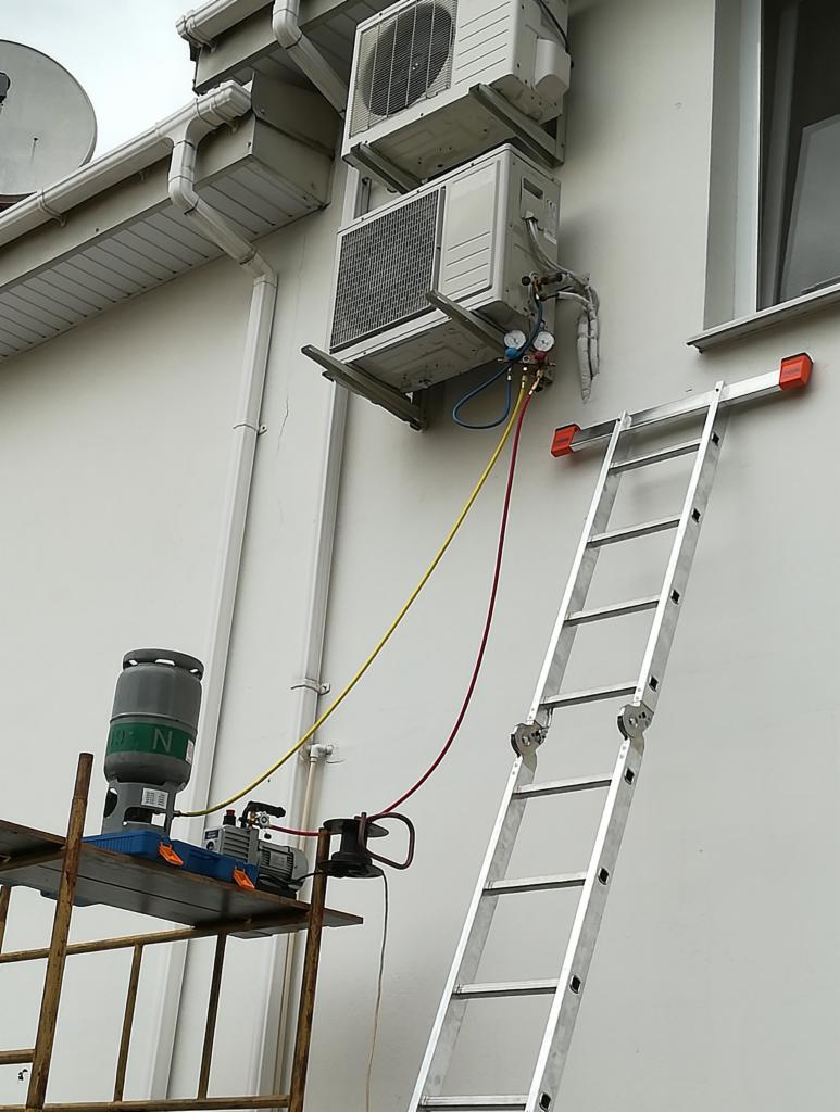 Dwa agregaty na ścianie budynku, podłączone do manometrów i butli z gazem, drabina oparta o ścianę, proces serwisowania.