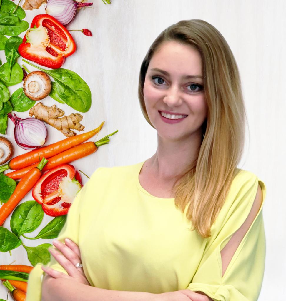 Uśmiechnięta blondynka w żółtej bluzce na tle z warzywami: marchew, papryka, szpinak, pieczarki, imbir i czerwona cebula.