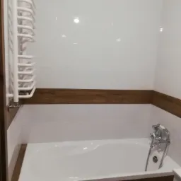 Biała wanna obudowana płytkami imitującymi drewno, z chromowaną armaturą i grzejnikiem drabinkowym na tle białych płytek ściennych z poziomym pasem dekoracyjnym.