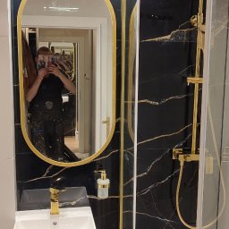 Rafał Witkowski - Wykończona łazienka z czarnymi płytkami imitującymi marmur ze złotymi żyłami, owalne lustro w złotej ramie, biała umywalka nablatowa z złotą armaturą, kabina prysznicowa ze złotymi elementami...