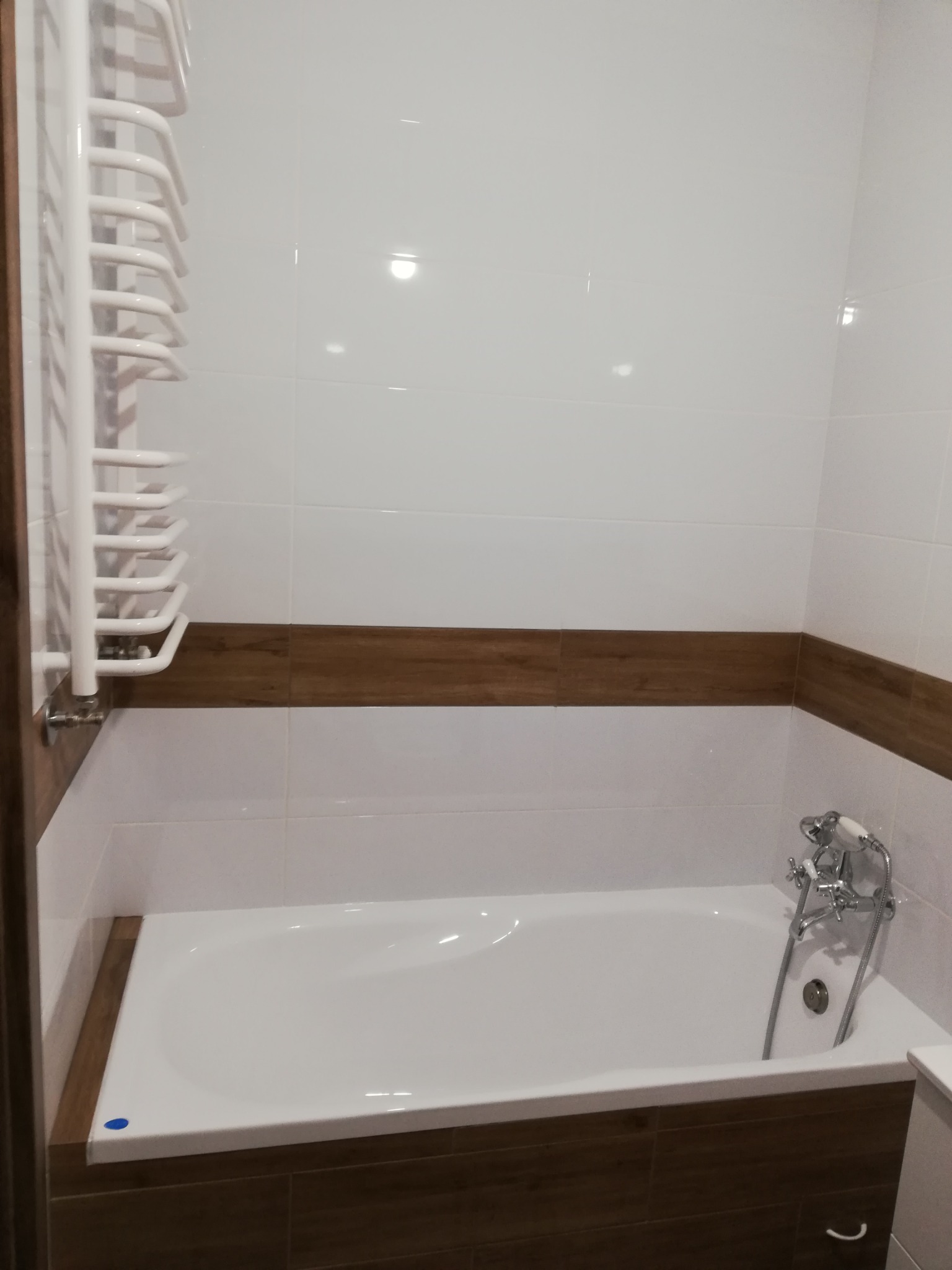 Biała wanna obudowana płytkami imitującymi drewno, z chromowaną armaturą i grzejnikiem drabinkowym na tle białych płytek ściennych z poziomym pasem dekoracyjnym.