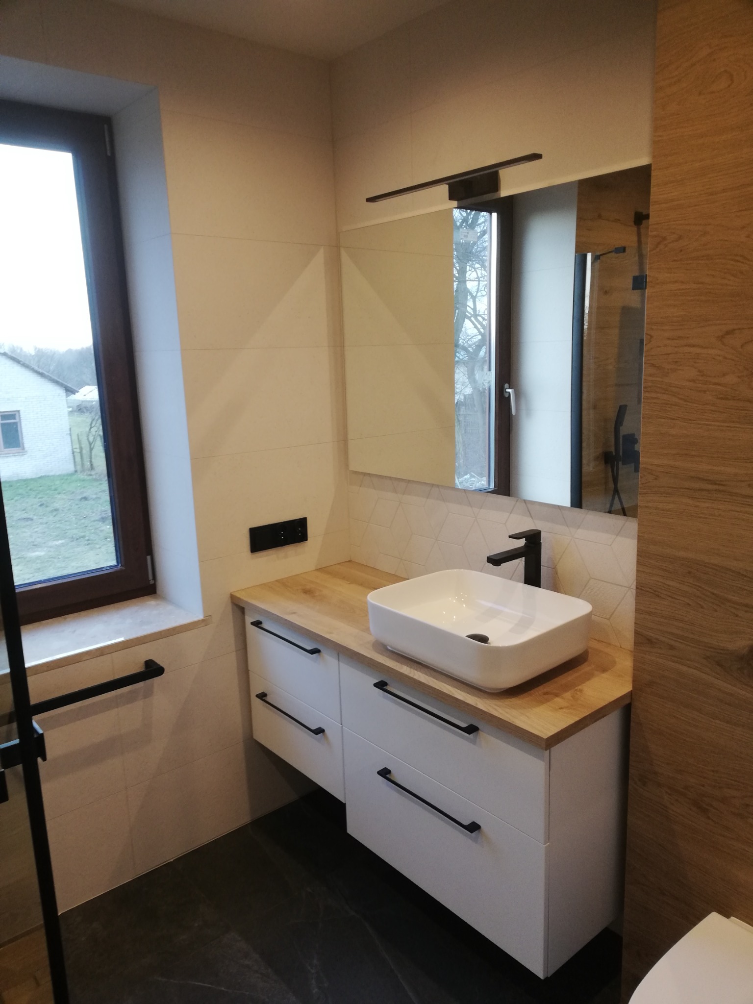 Nowoczesna łazienka z białą szafką pod umywalkę, drewnianym blatem, czarną armaturą i lustrem z oświetleniem LED. Widoczne jasne płytki na ścianach i ciemna podłoga z imitacją kamienia. Za oknem...