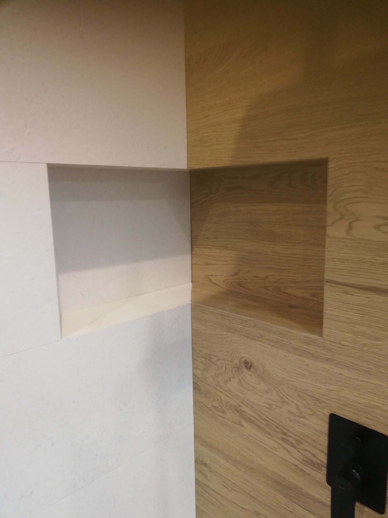 Narożnik wnętrza z wnęką w ścianie wyłożonej białymi płytami oraz fragmentem ściany z okładziną drewnopodobną i czarnym uchwytem.