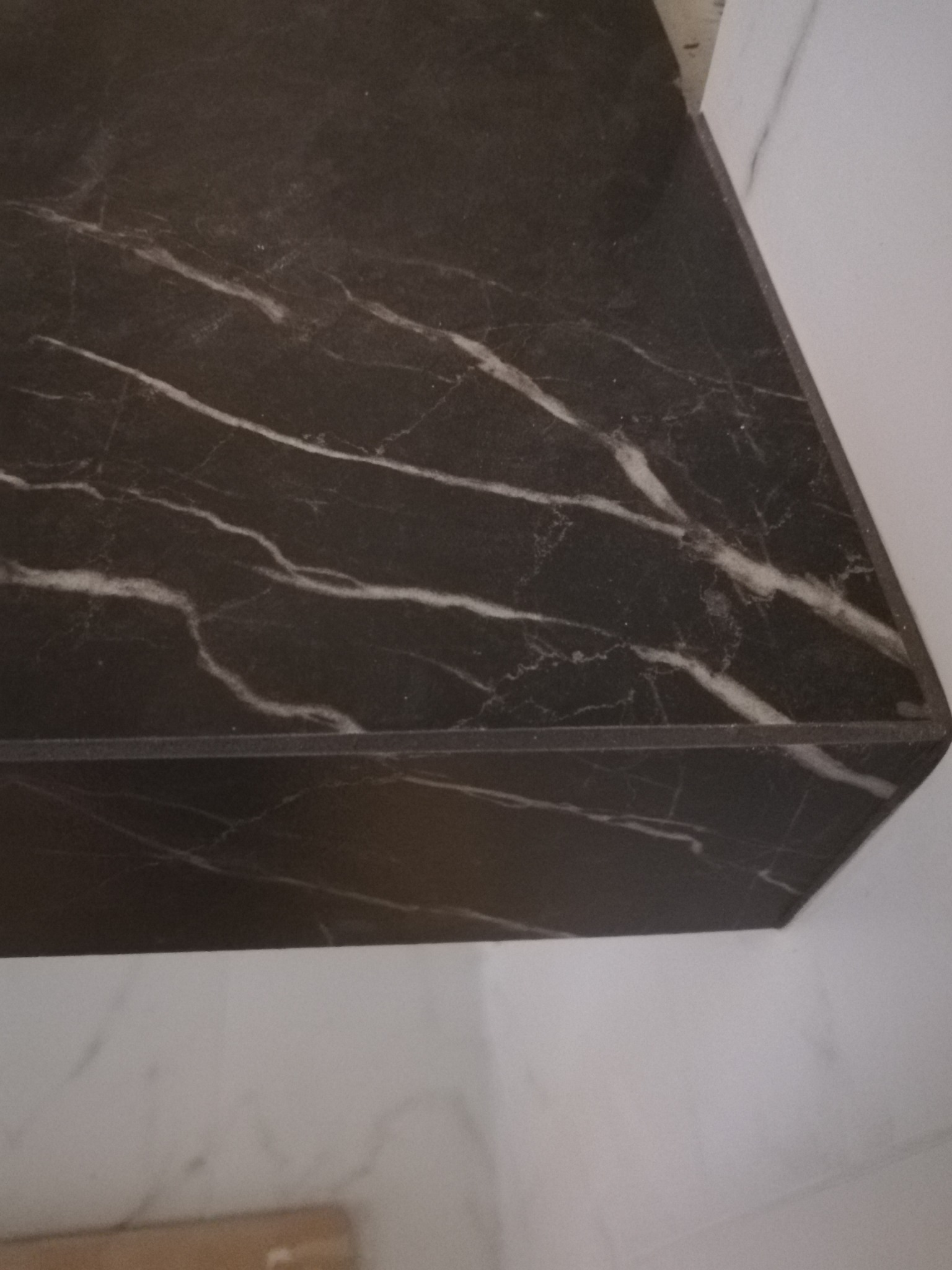 Fragment blatu z ciemnego marmuru z białymi żyłami, przymocowanego do ściany wykończonej białymi płytkami marmuropodobnymi.