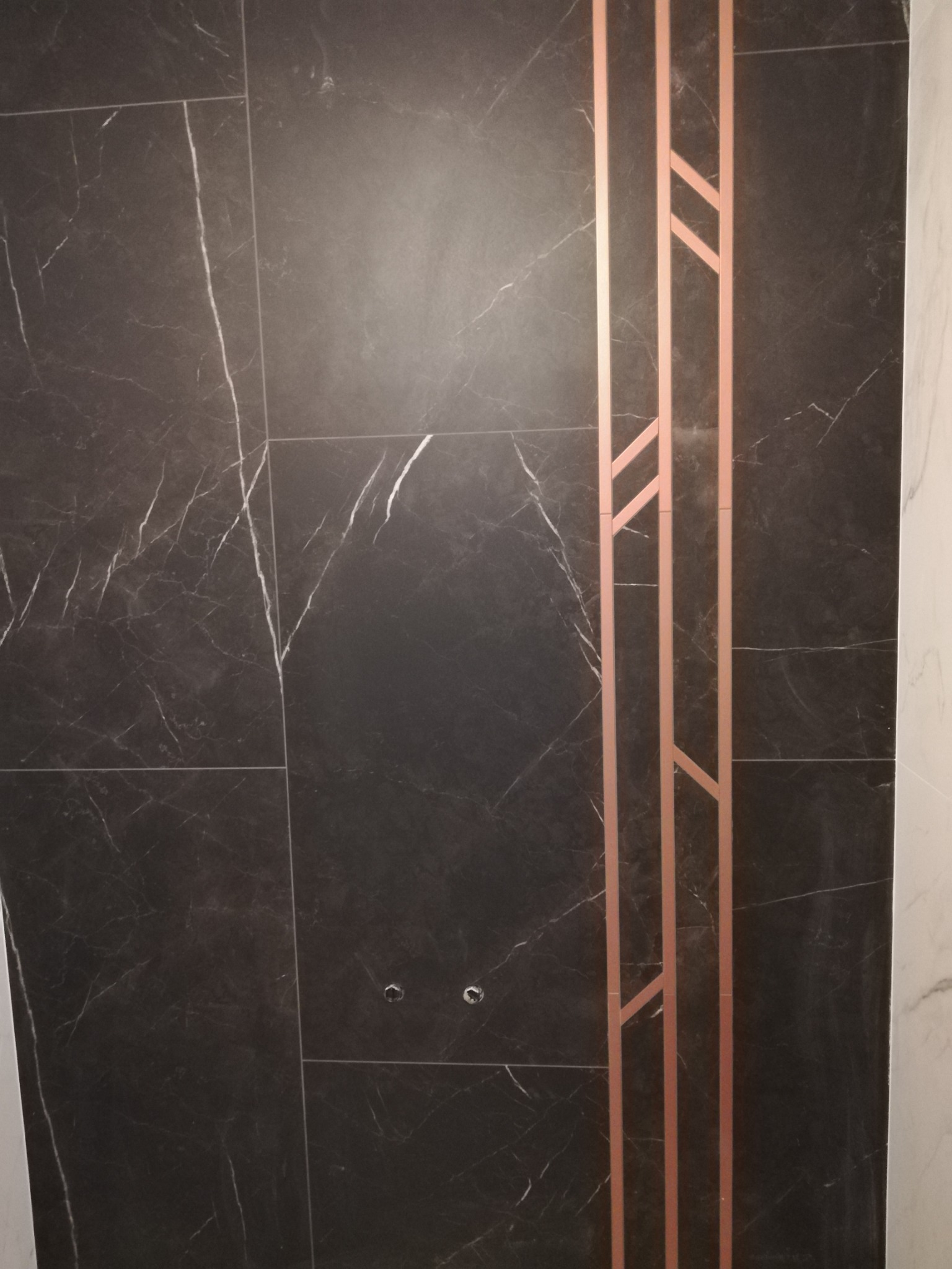 Ściana wyłożona czarnymi płytkami imitującymi marmur z dekoracyjnym, geometrycznym wzorem z miedzianych listew.
