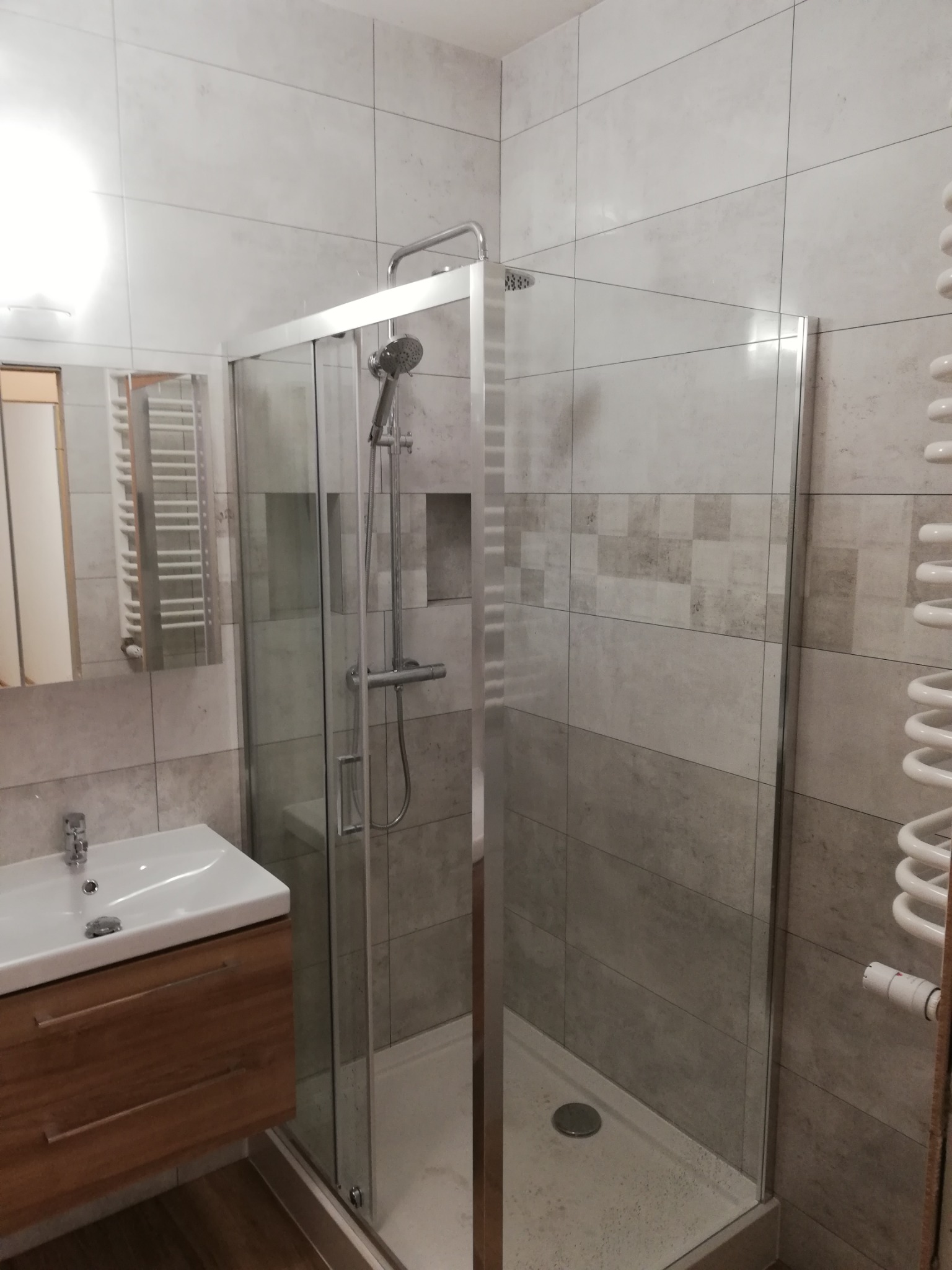 Nowoczesna łazienka z kabiną prysznicową ze szklanymi drzwiami przesuwnymi, umywalką nablatową z szafką z drewnopodobnego materiału oraz grzejnikiem drabinkowym na ścianie wyłożonej płytkami...