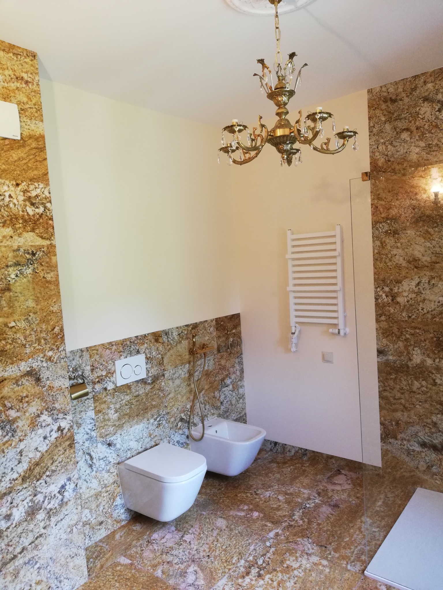 Elegancka łazienka z marmurowymi płytkami w odcieniach brązu i beżu, wyposażona w białą armaturę, grzejnik drabinkowy i ozdobny, złoty żyrandol.