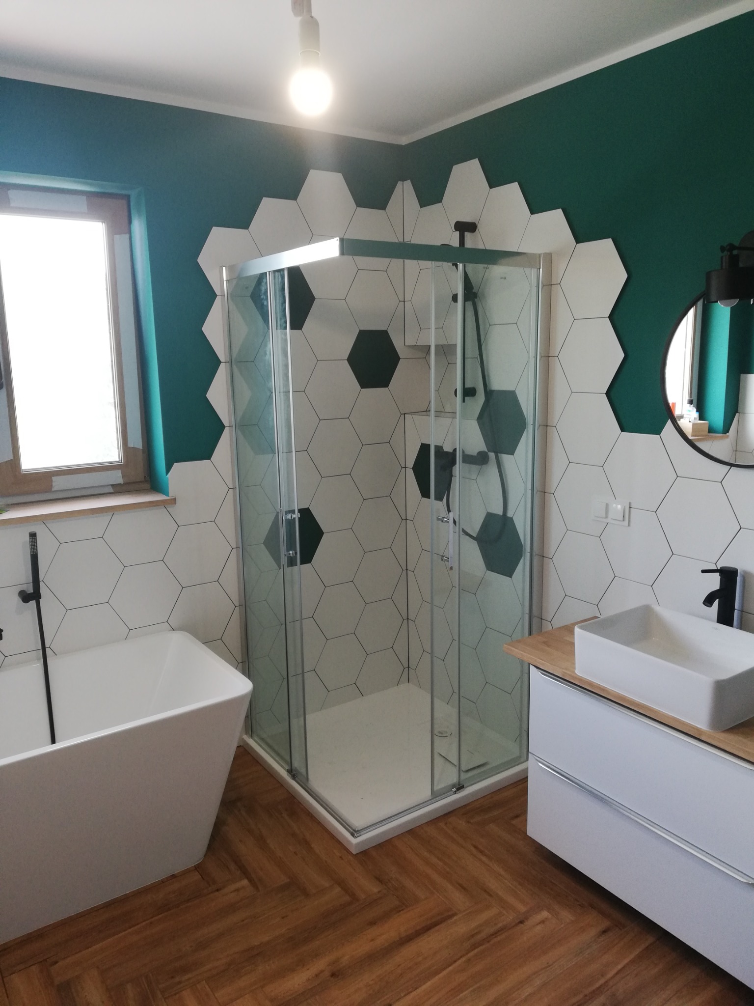 Nowoczesna łazienka z kabiną prysznicową w rogu, białą wanną wolnostojącą i umywalką nablatową na szafce z drewnianym blatem, ściany wykończone heksagonalnymi płytkami w kolorze białym i zielonym...