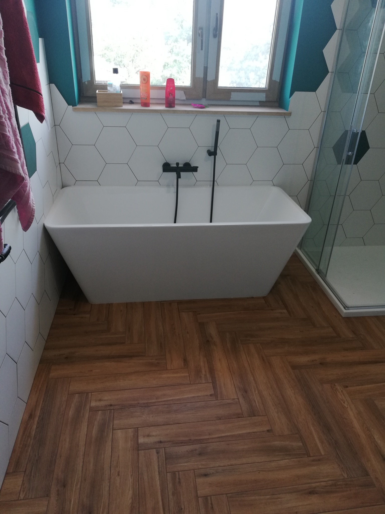 Nowoczesna łazienka z białą, prostokątną wanną, heksagonalnymi płytkami na ścianach, drewnopodobną podłogą ułożoną w jodełkę i kabiną prysznicową w rogu.