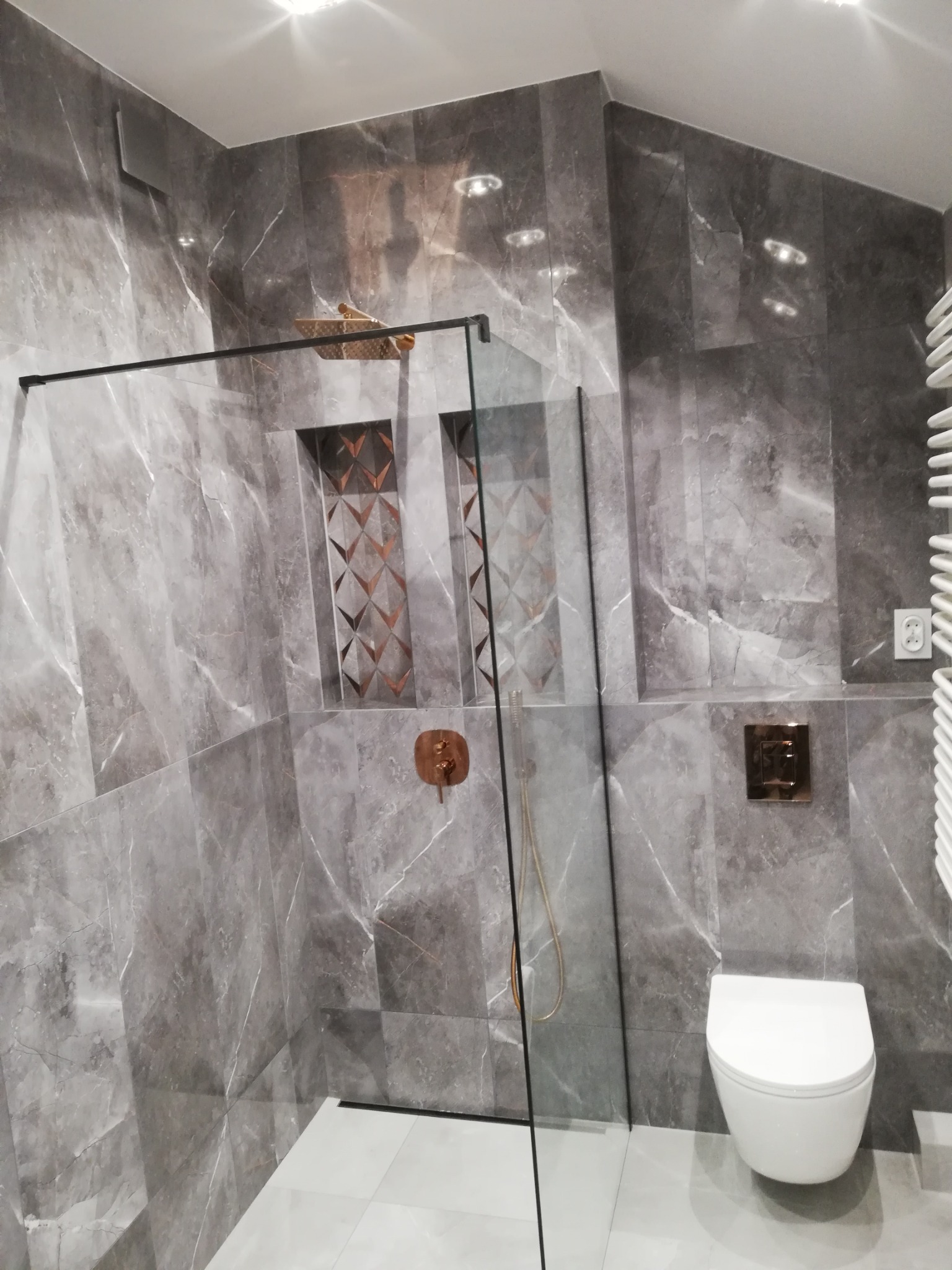 Elegancka łazienka w Legionowie z szarymi płytkami imitującymi marmur, kabiną prysznicową typu walk-in i dekoracyjnymi wnękami wykończonymi mozaiką w odcieniach miedzi.