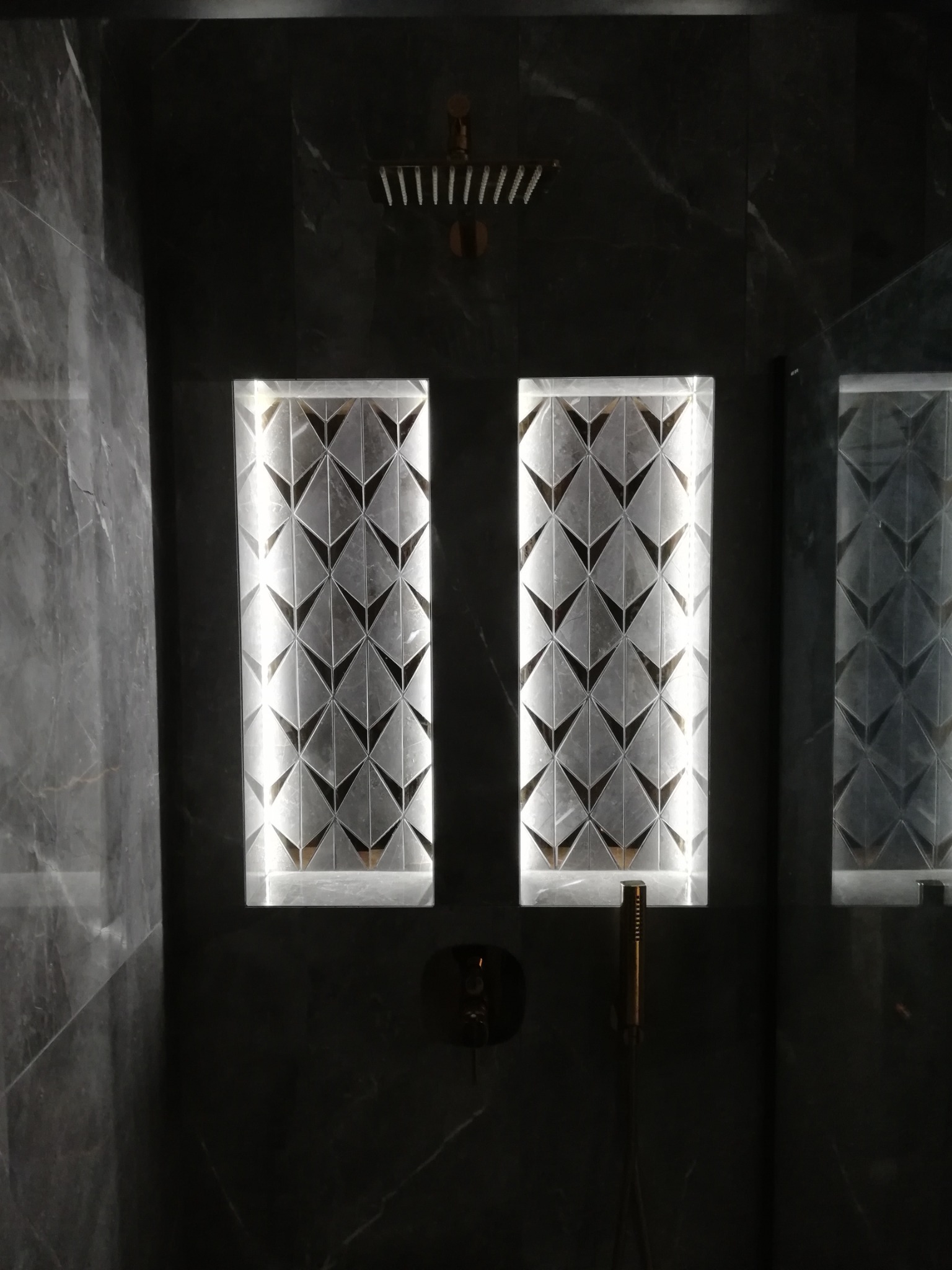 Nowoczesna łazienka z ciemnym marmurem, podświetlane nisze z geometrycznym wzorem i deszczownica w kolorze miedzi.