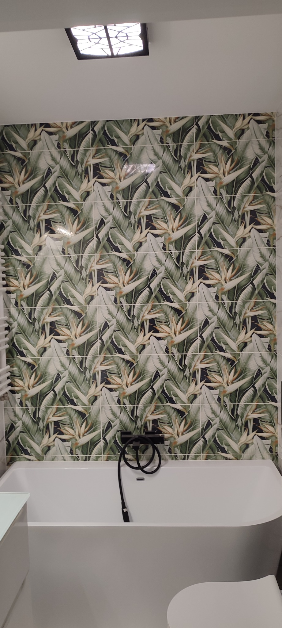 Biała wanna wolnostojąca z czarną baterią na tle ściany wyłożonej płytkami z motywem tropikalnych liści i kwiatów strelicji, oświetlenie sufitowe w czarnej oprawie.