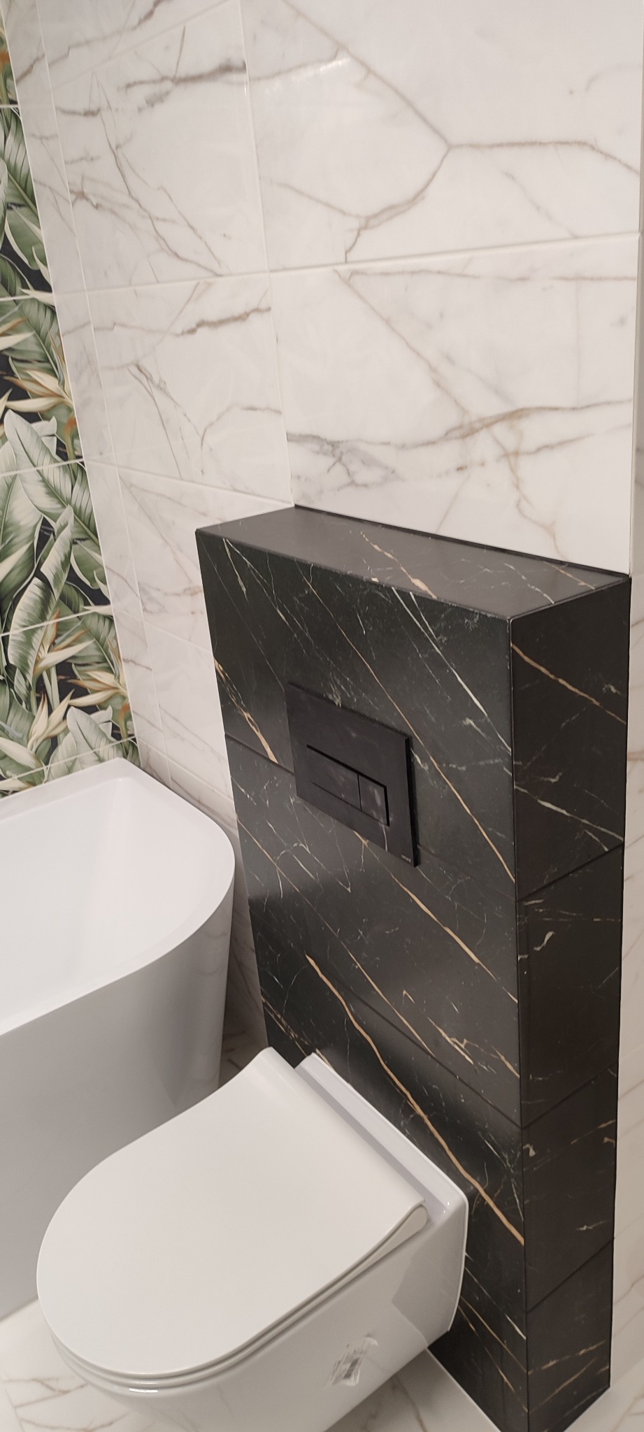 Nowoczesna toaleta podwieszana z czarną obudową ze złotymi żyłkami, pasująca do czarnego przycisku spłukującego, na tle białych płytek z marmurkowym wzorem i fragmentu wanny oraz ściany z motywem...