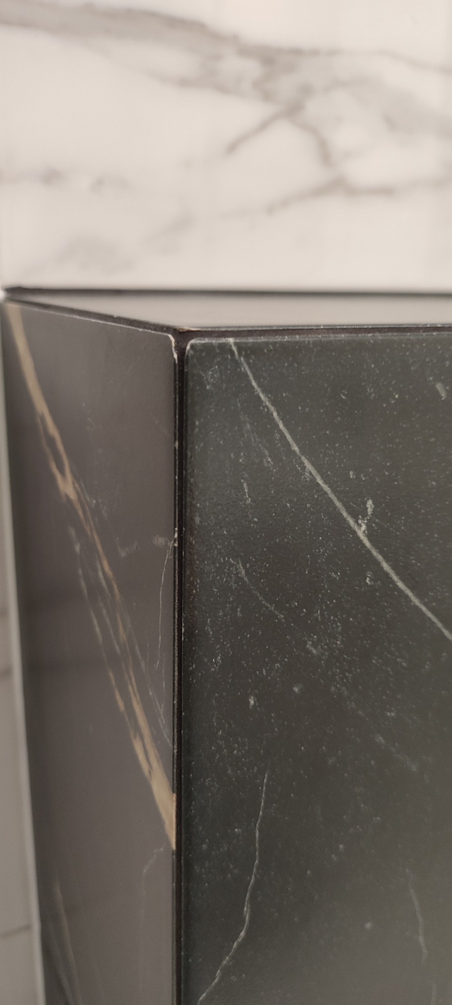 Fragment narożnika obudowy z płytek imitujących czarny marmur z widocznymi żyłami, na tle białej ściany z marmurowym wzorem, ukazujący precyzję łączenia elementów.