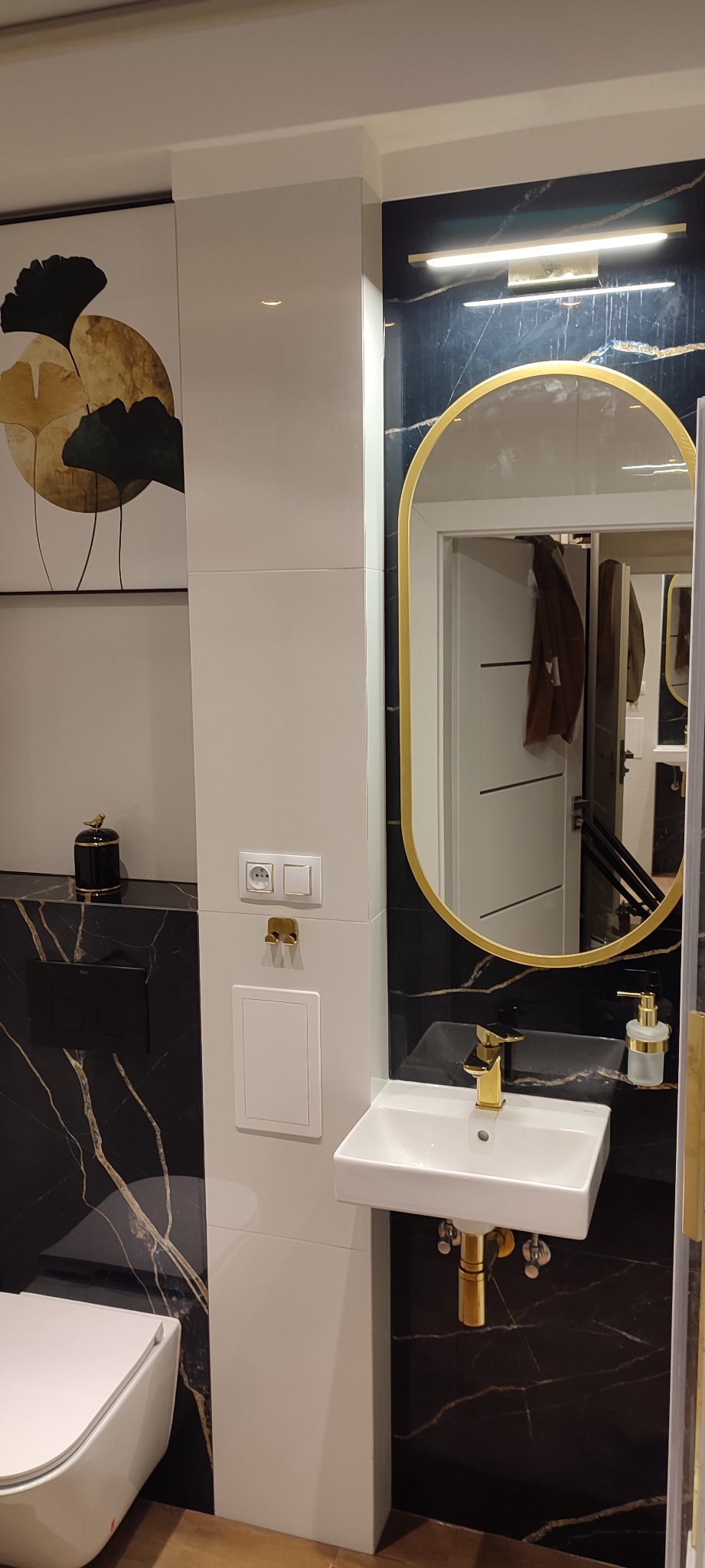 Nowoczesna łazienka z czarnymi marmurowymi płytkami, białymi ścianami i złotymi akcentami, w tym owalne lustro, bateria umywalkowa i dozownik mydła.