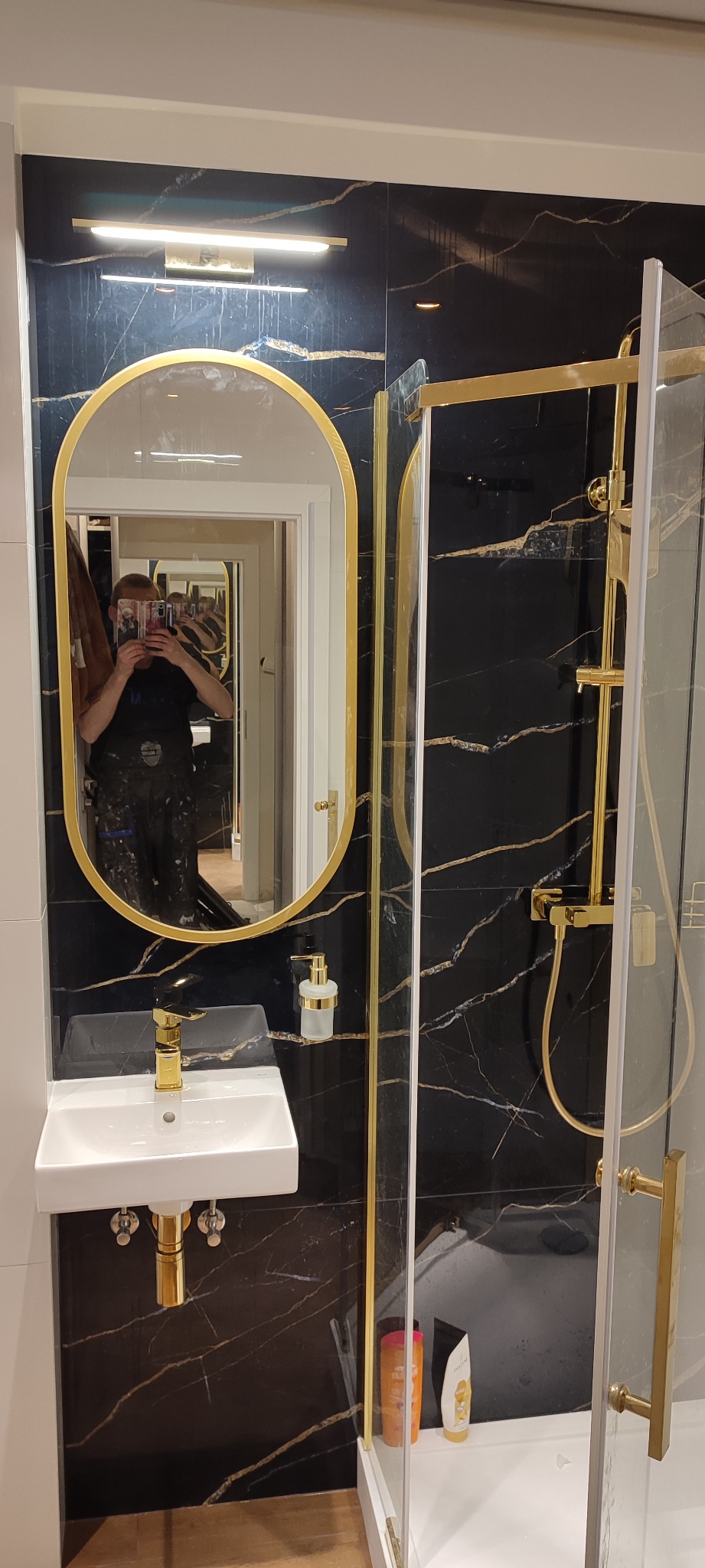 Wykończona łazienka z czarnymi płytkami imitującymi marmur ze złotymi żyłami, owalne lustro w złotej ramie, biała umywalka nablatowa z złotą armaturą, kabina prysznicowa ze złotymi elementami...