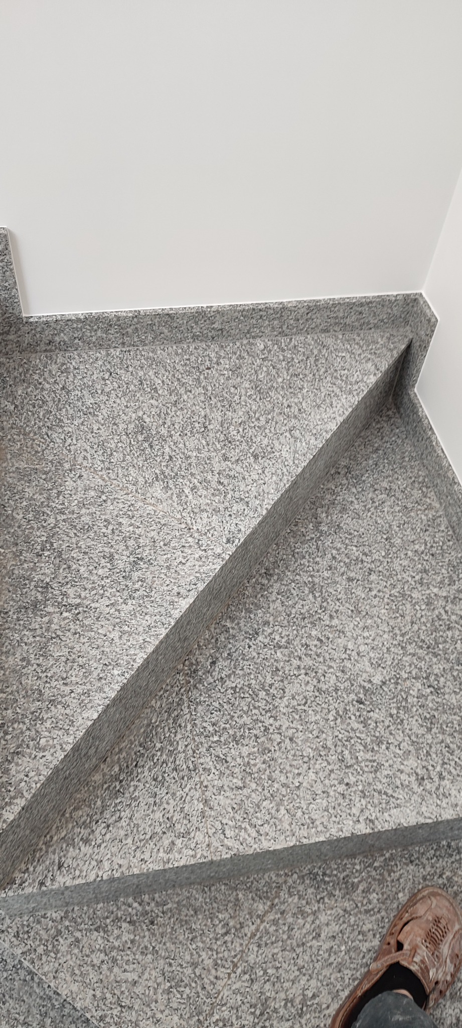 Granitowe schody wykończone z precyzją, widoczne z góry, z fragmentem buta w prawym dolnym rogu, kontrastujące z białą ścianą w tle.