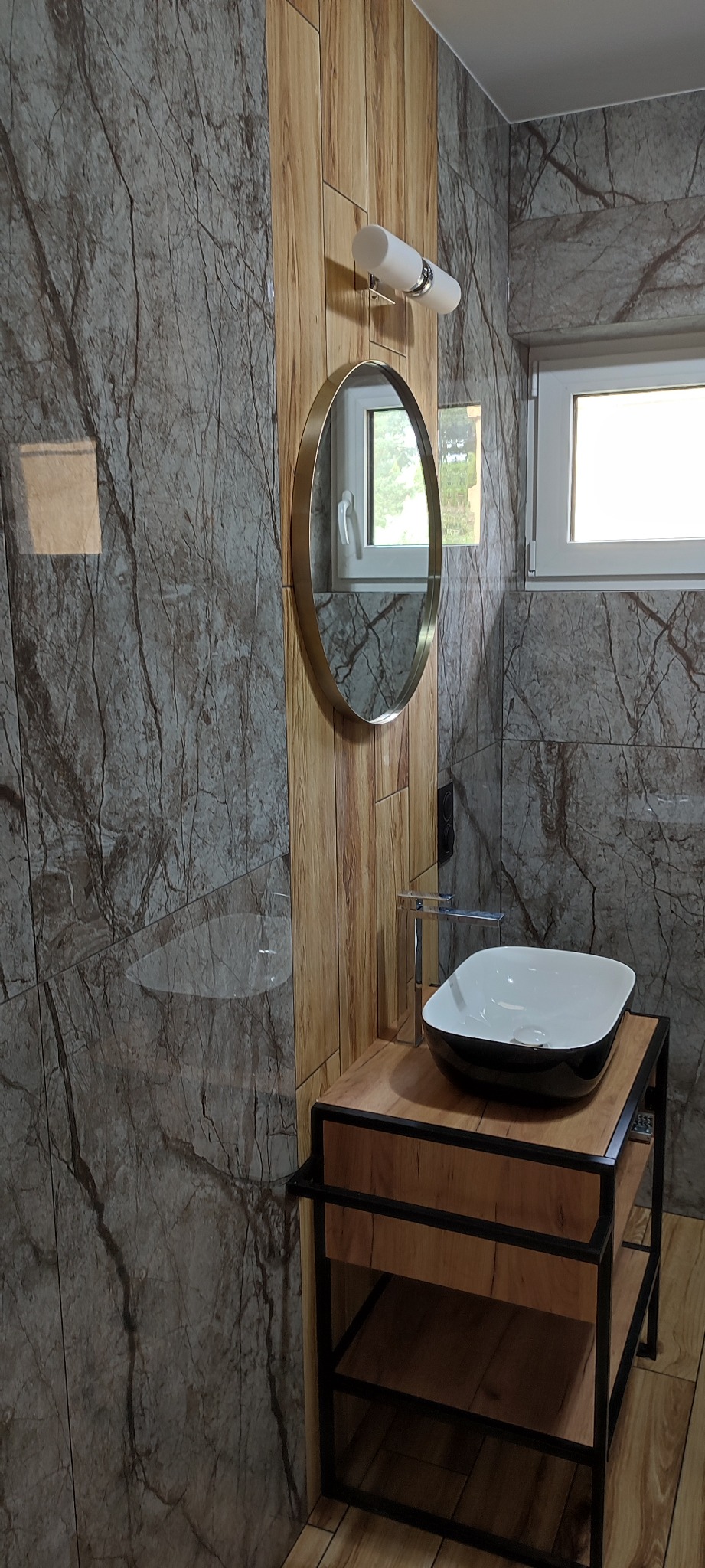 Nowoczesna łazienka z umywalką nablatową w czarno-białym kolorze, owalnym lustrem w złotej ramie i płytkami imitującymi marmur oraz drewno.