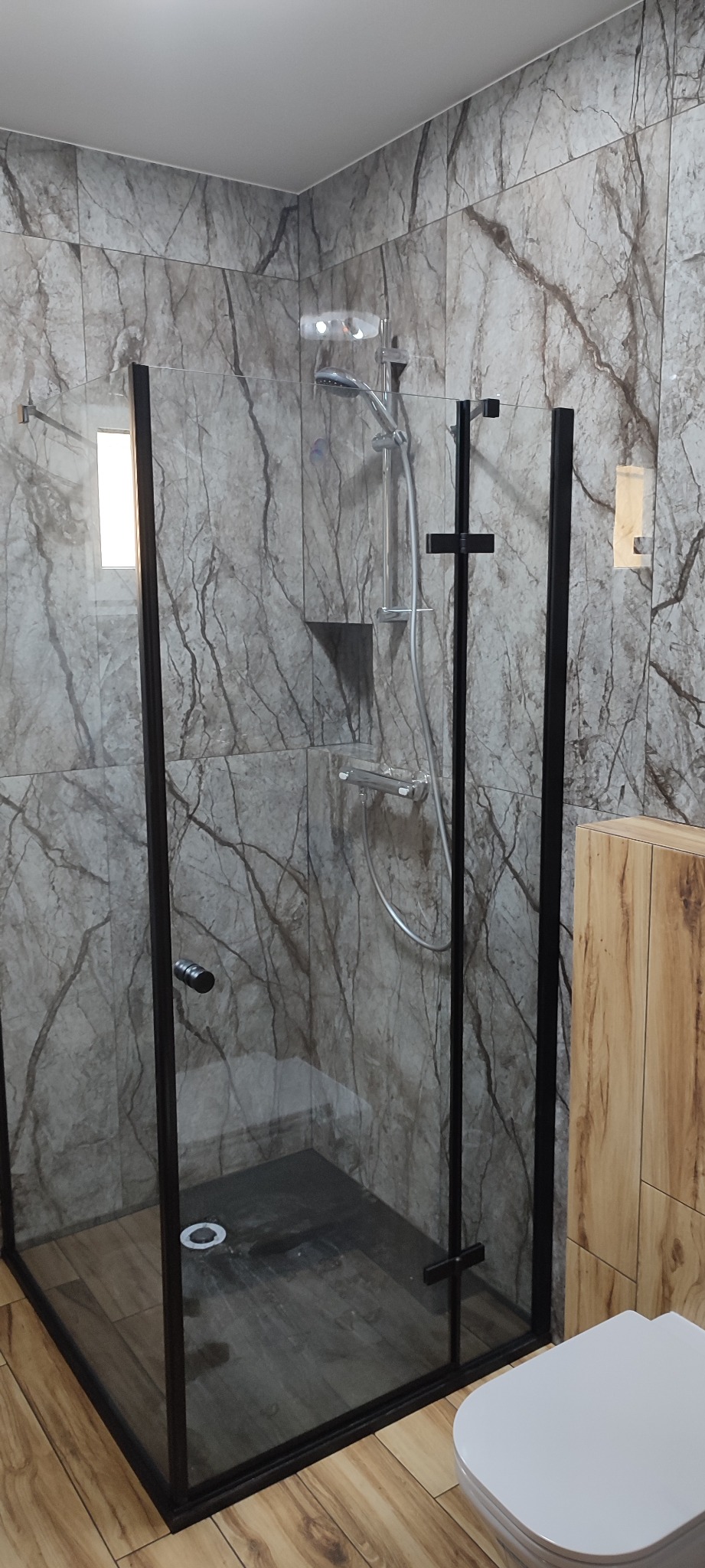 Nowoczesna kabina prysznicowa z czarnymi ramami i szklanymi ścianami, wykończona płytkami imitującymi marmur, z widocznym odpływem liniowym i częścią toalety w rogu.