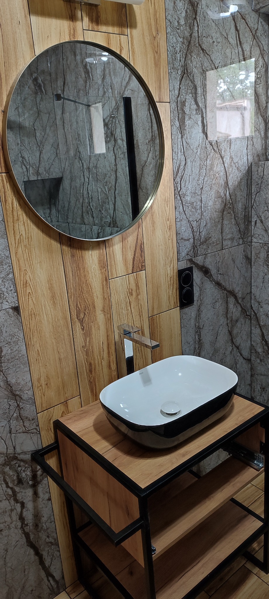Nowoczesna łazienka z okrągłym lustrem w złotej ramie, umywalką nablatową w kolorze biało-czarnym oraz baterią chromowaną. Ściany wyłożone płytkami imitującymi drewno i marmur. Umywalka osadzona...
