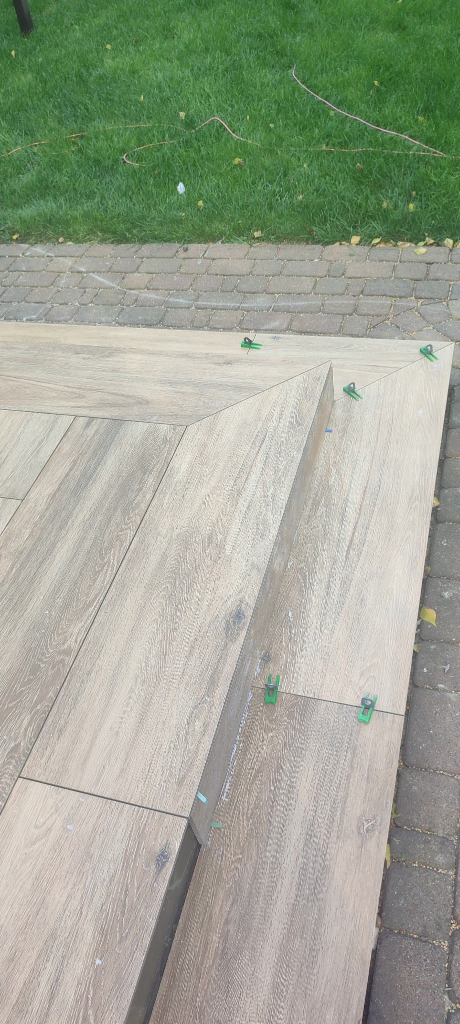 Układanie płytek imitujących drewno na zewnątrz, widoczne zielone klipsy dystansowe, trawnik w tle.