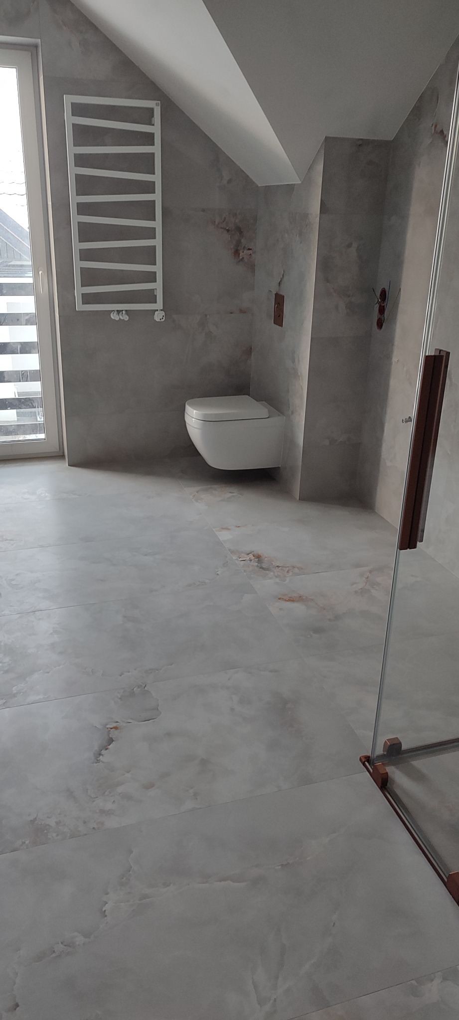 Wykończona łazienka na poddaszu z podwieszaną toaletą, grzejnikiem drabinkowym i szklaną kabiną prysznicową, wykończona płytkami imitującymi marmur.