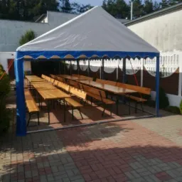 Biały namiot z niebieskimi akcentami, rozstawiony na zewnątrz, z drewnianymi stołami i ławkami w środku, przygotowany na imprezę plenerową.