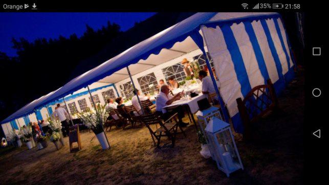 Uroczystość w namiocie z białymi i niebieskimi pasami, oświetlonym lampionami i udekorowanym kwiatami, w tle ciemny las.