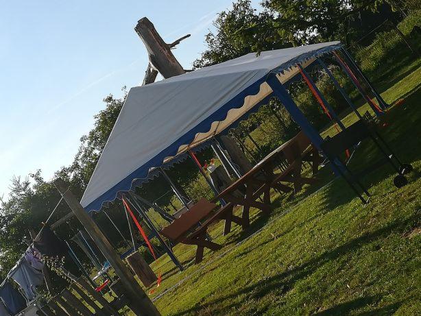 Namiot imprezowy z niebieskimi falbanami w Bojano, z drewnianymi stołami i ławkami wewnątrz, ustawiony na trawie obok pnia drzewa.