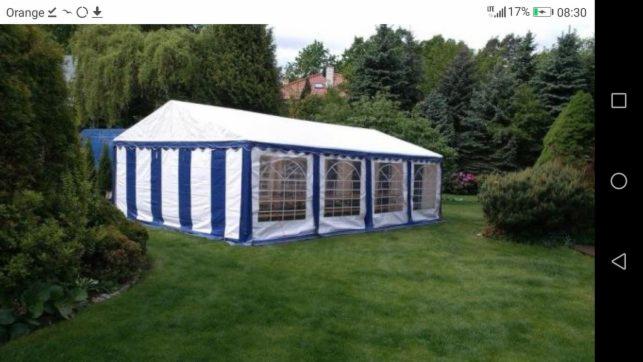 Rozłożony biały namiot z niebieskimi pasami na trawniku, otoczony zielenią drzew i krzewów, widok z boku.