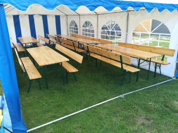 Wnętrze namiotu imprezowego z drewnianymi stołami i ławkami na trawie, widok z boku, niebieskie wykończenie dachu namiotu.