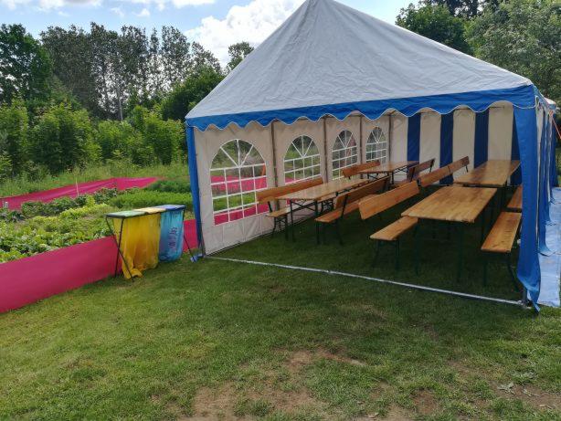 Namiot imprezowy z niebieskimi akcentami, ustawiony na trawie obok upraw warzyw, z widocznymi stołami i ławkami wewnątrz oraz koszami na śmieci na zewnątrz.
