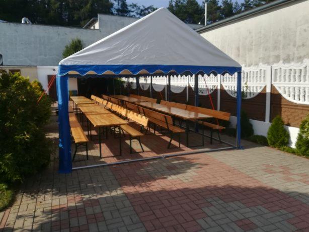 Biały namiot z niebieskimi akcentami, rozstawiony na zewnątrz, z drewnianymi stołami i ławkami w środku, przygotowany na imprezę plenerową.