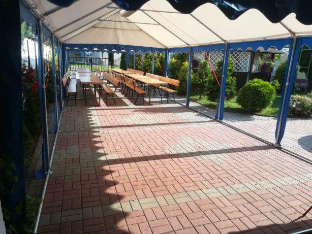 Przestronny namiot z niebieskimi akcentami, ustawiony na ceglanym patio, z widocznymi drewnianymi stołami i ławkami, otoczony zielenią.