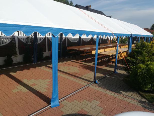 Długi, biały namiot z niebieskim obramowaniem, rozstawiony na ceglanym patio z drewnianymi stołami i ławkami wewnątrz, oświetlony popołudniowym słońcem.